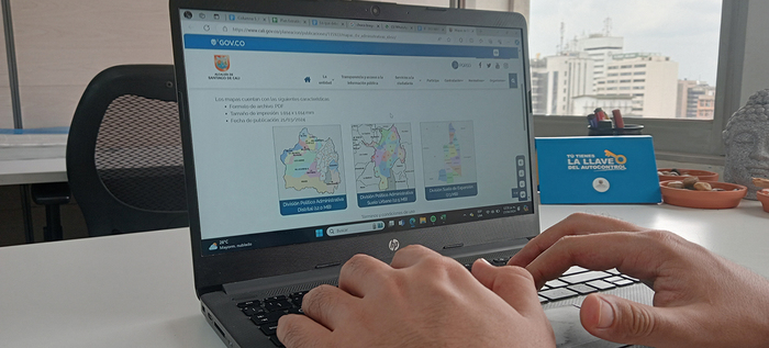 Fotográfía en un puesto de trabajo de la oficina del Departamento Administrativo de Planeación, donde se muestra un computador portátil y unas manos sobre el teclado, y en la pantalla la publicación de los mapas de la Diviisón Político Administrativa de Santiago de Cali.